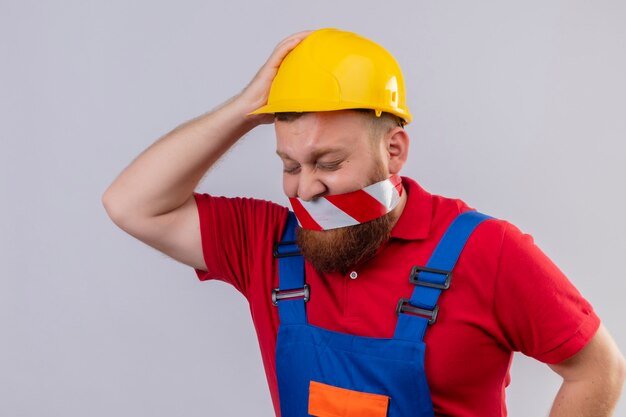 Jeune homme constructeur barbu en uniforme de construction et casque de sécurité avec du ruban adhésif sur sa bouche à la confusion et déçu avec la main sur sa tête pour erreur