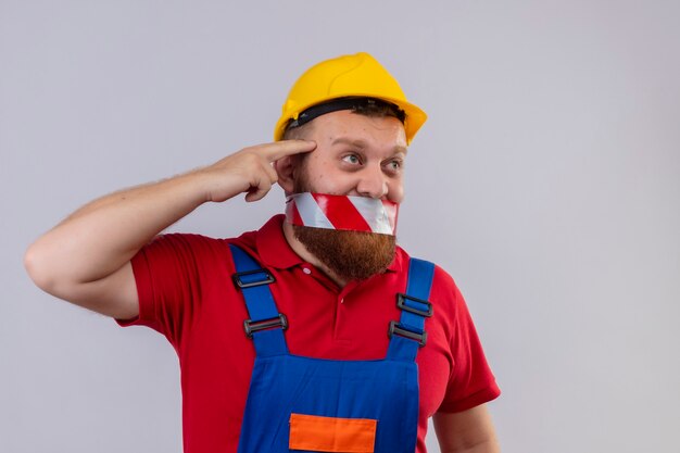 Jeune homme constructeur barbu en uniforme de construction et casque de sécurité avec du ruban adhésif sur sa bouche à côté confus en pointant sa tempe