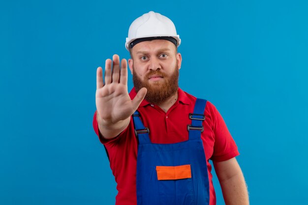 Jeune homme constructeur barbu en uniforme de construction et casque de sécurité avec main ouverte faisant panneau d'arrêt avec visage sérieux