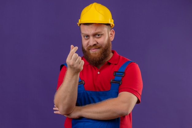 Jeune Homme Constructeur Barbu En Uniforme De Construction Et Casque De Sécurité Mécontent De Faire Le Geste De L'argent Avec La Main Frottant Les Doigts Demandant De L'argent Sur Backgroun Violet