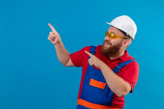 Jeune homme de constructeur barbu en uniforme de construction et casque de sécurité souriant heureux et positif pointant avec les doigts sur le côté