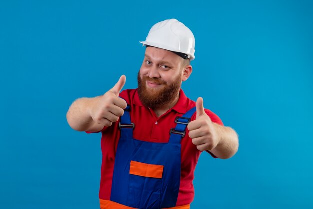 Jeune homme constructeur barbu en uniforme de construction et casque de sécurité souriant sympathique montrant les pouces vers le haut avec les deux mains