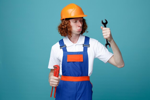 Jeune homme constructeur confus en uniforme tenant et regardant des clés isolées sur fond bleu