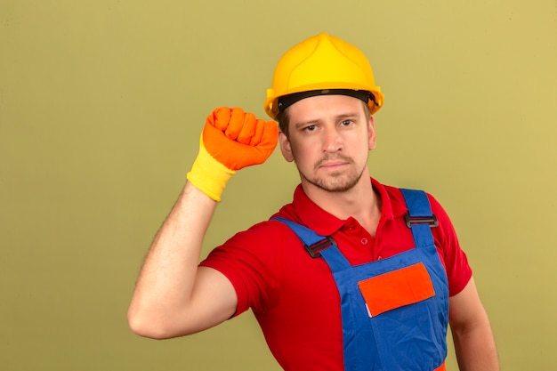 Jeune homme constructeur en construction gants uniformes et casque de sécurité en levant le poing avec concept gagnant visage sérieux sur mur vert isolé