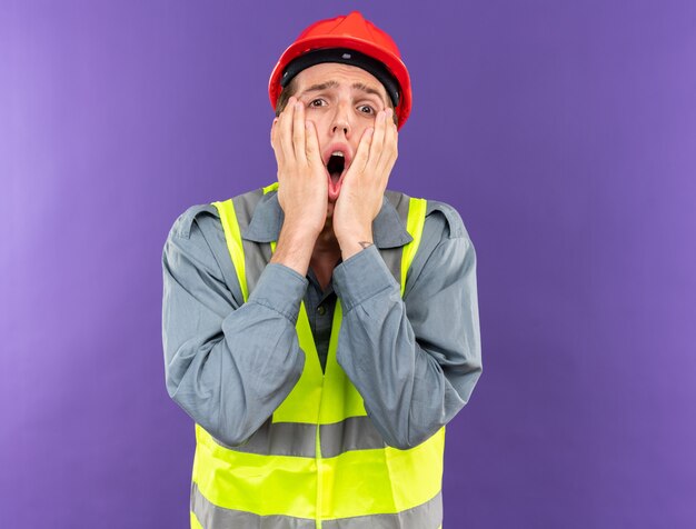 Jeune homme de constructeur effrayé en uniforme mettant les mains sur les joues