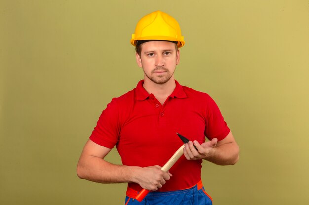 Jeune homme constructeur en polo rouge et casque de sécurité tenant un marteau avec un visage sérieux sur mur vert isolé