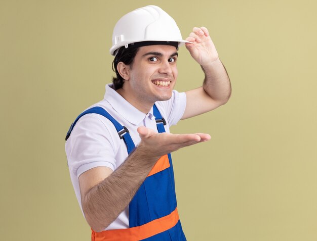 Jeune homme constructeur en uniforme de construction et casque de sécurité à l'avant avec le bras en souriant joyeusement debout sur le mur vert