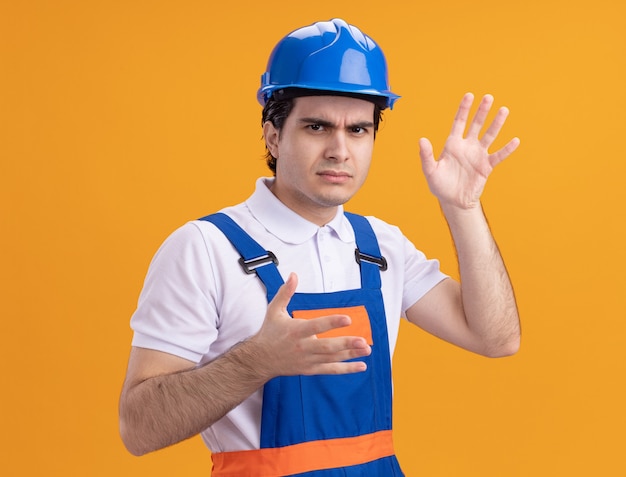 Jeune homme constructeur en uniforme de construction et casque de sécurité à l'avant d'être mécontent avec les bras debout sur le mur orange