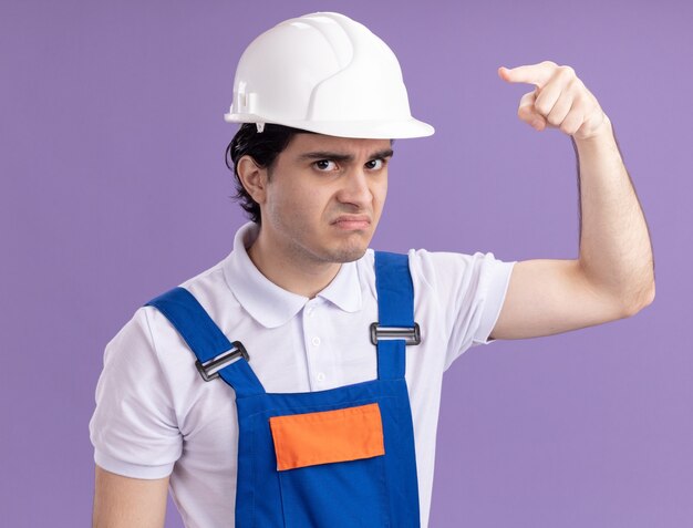 Jeune homme constructeur en uniforme de construction et casque de sécurité à l'avant avec le visage en colère pointant avec l'index à l'avant debout sur le mur violet