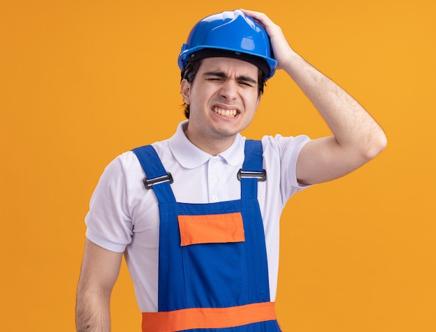 Jeune homme constructeur en uniforme de construction et casque de sécurité à la confusion et très anxieux avec la main sur la tête pour erreur debout sur un mur orange