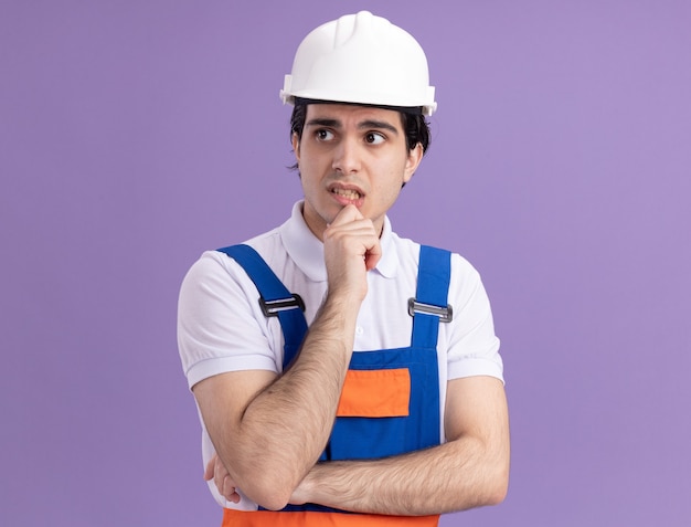 Photo gratuite jeune homme constructeur en uniforme de construction et casque de sécurité à côté avec une expression pensive pensant debout sur un mur violet