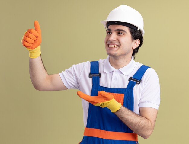 Jeune homme constructeur en uniforme de construction et casque de sécurité dans des gants en caoutchouc à côté montrant les pouces vers le haut souriant joyeusement debout sur le mur vert