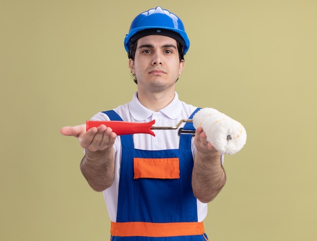 Jeune homme constructeur en uniforme de construction et casque de sécurité démontrant rouleau à peinture à l'avant avec un visage sérieux debout sur un mur vert