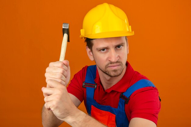 Jeune homme constructeur en uniforme de construction et casque de sécurité avec une expression de colère menaçant de frapper avec un marteau sur un mur orange isolé