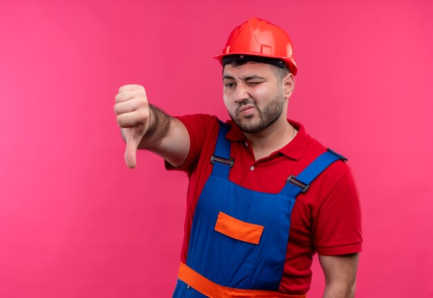 Jeune homme constructeur en uniforme de construction et casque de sécurité montrant les pouces vers le bas mécontent