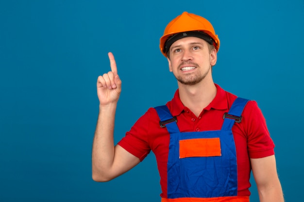 Jeune homme constructeur en uniforme de construction et casque de sécurité pointant avec le doigt vers le haut avec le sourire sur le visage face au mur bleu isolé