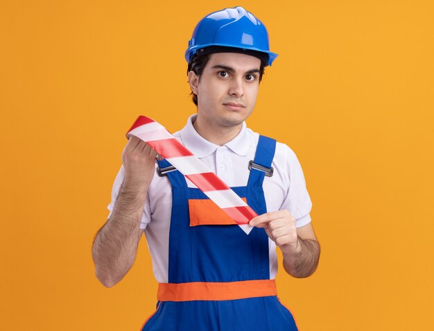 Jeune homme constructeur en uniforme de construction et casque de sécurité tenant une bande d'avertissement à l'avant avec une expression sérieuse et confiante debout sur un mur orange