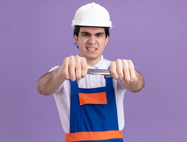 Jeune homme constructeur en uniforme de construction et casque de sécurité tenant la clé en le regardant avec le visage en colère debout sur le mur violet