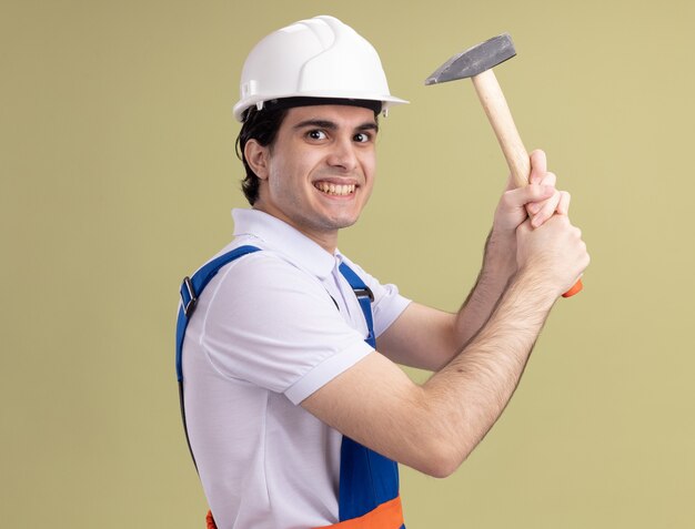 Jeune homme constructeur en uniforme de construction et casque de sécurité tenant un marteau à l'avant souriant sournoisement debout sur un mur vert