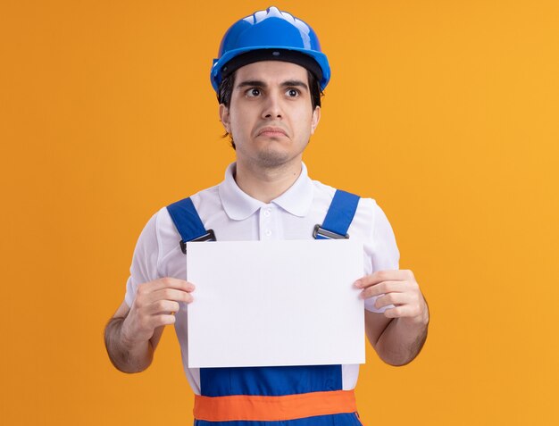 Jeune homme constructeur en uniforme de construction et casque de sécurité tenant une page blanche à côté avec une expression triste sur le visage debout sur un mur orange