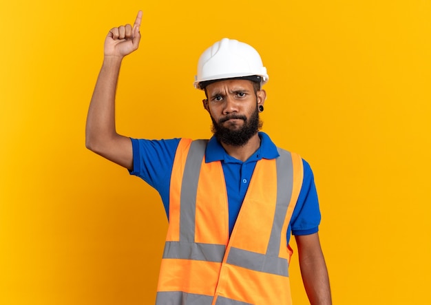 Jeune homme de construction afro-américain mécontent en uniforme avec un casque de sécurité pointant vers le haut isolé sur fond orange avec espace de copie