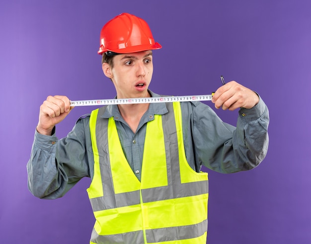 Jeune Homme De Construction Concerné En Uniforme S'étendant Sur Un Ruban à Mesurer Isolé Sur Un Mur Bleu