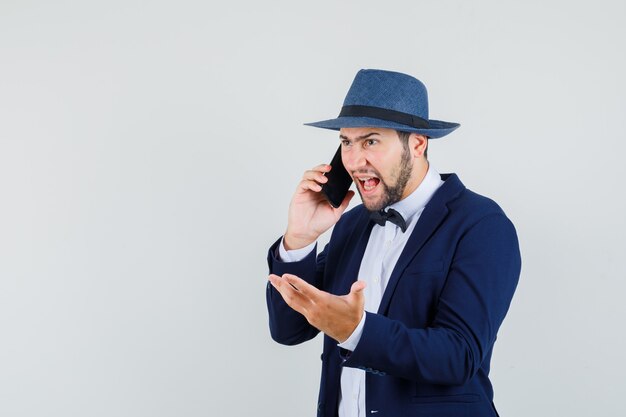 Jeune homme en costume, chapeau criant sur téléphone mobile et à la furieuse, vue de face.