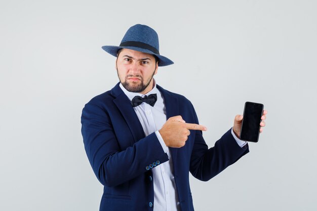 Jeune homme en costume, chapeau pointant sur le téléphone mobile et à la recherche de mécontentement, vue de face.