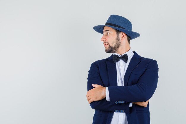 Jeune homme en costume, chapeau regardant de côté avec les bras croisés et à la recherche élégante, vue de face.