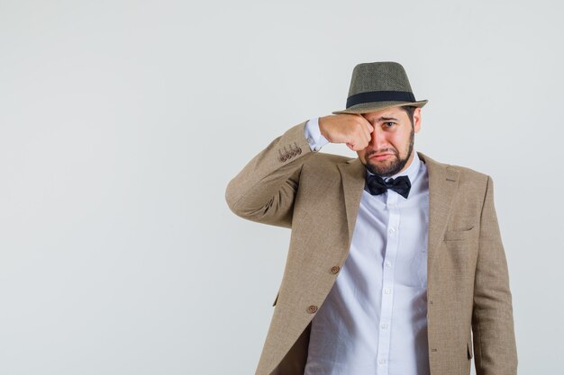 Jeune homme en costume, chapeau se frottant les yeux en pleurant comme un enfant et à la vue offensée, de face.