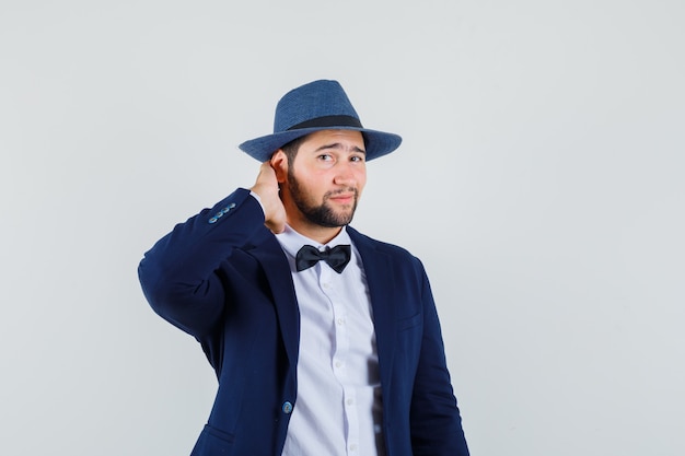 Jeune homme en costume, chapeau tenant la main sur le cou et regardant beau, vue de face.