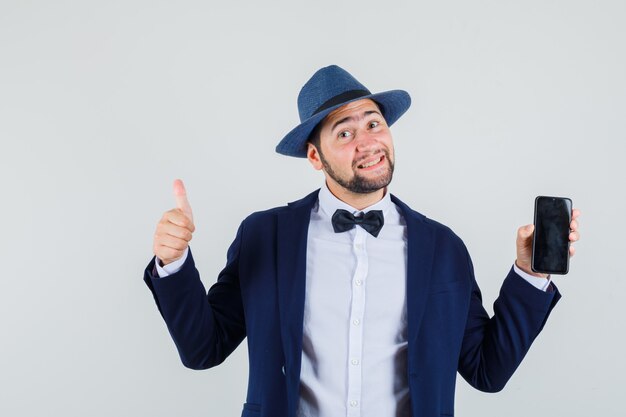 Jeune homme en costume, chapeau tenant le téléphone mobile avec le pouce vers le haut et à la joyeuse vue de face.