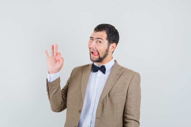 Jeune homme en costume montrant le geste ok et qui sort la langue, vue de face.