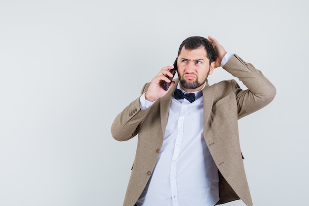 Jeune homme en costume parlant au téléphone mobile et à la vue de face, hésitant.