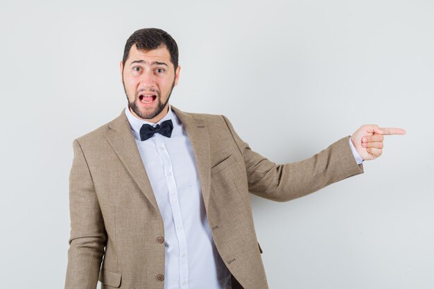 Jeune homme en costume pointant vers le côté et regardant anxieux, vue de face.