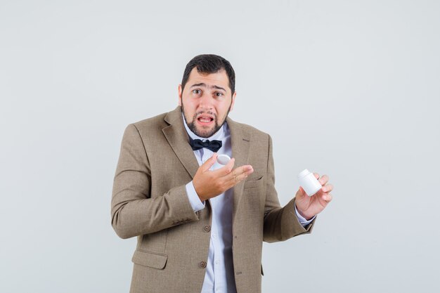 Jeune homme en costume tenant une bouteille vide de pilules et regardant impuissant, vue de face.