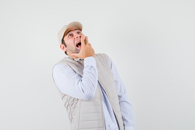 Jeune homme couvrant la bouche avec la main, regardant vers le haut en veste beige et casquette et à la recherche de plaisir. vue de face.