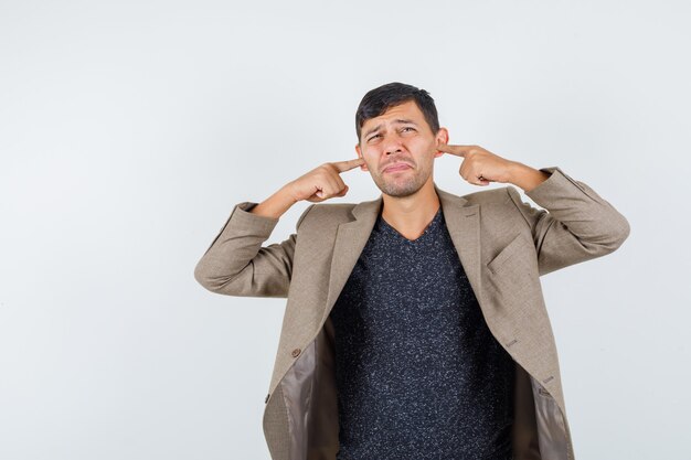 Jeune homme couvrant ses oreilles avec les doigts en veste marron grisâtre, chemise noire et à l'insatisfaction, vue de face.