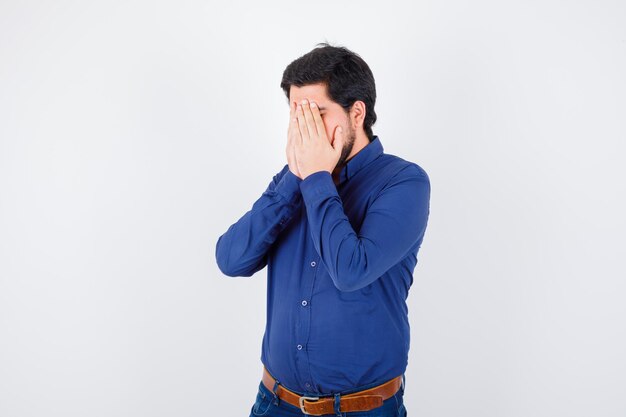 Jeune homme couvrant le visage avec les mains en chemise, jeans et l'air stressé. .