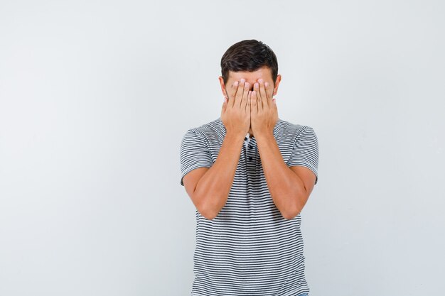 Jeune homme couvrant le visage avec les mains en t-shirt et l'air déprimé. vue de face.