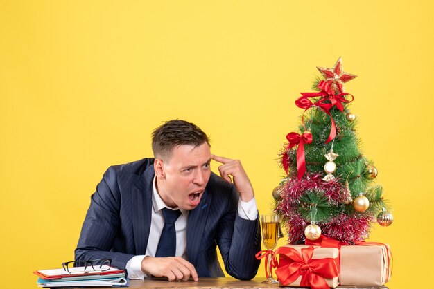 Jeune homme criant assis à la table près de l'arbre de Noël et des cadeaux sur jaune