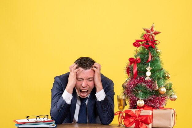 Jeune homme criant tout en tenant sa tête assise à la table près de l'arbre de Noël et présente sur jaune