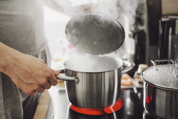 Jeune homme cuisinant des aliments frais à la maison et ouvrant le couvercle du pot fumant.