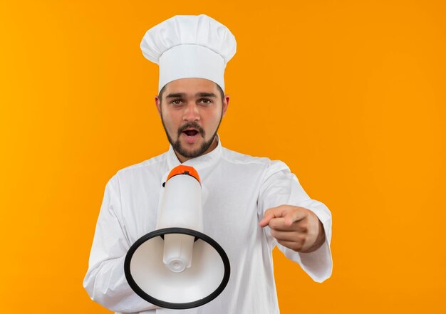 Photo gratuite jeune homme cuisinier impressionné en uniforme de chef parlant par le haut-parleur et pointant isolé sur un mur orange avec espace de copie