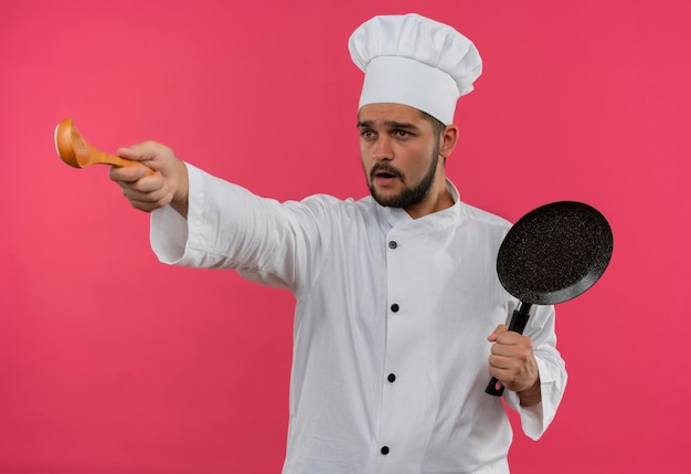 Jeune homme cuisinier impressionné en uniforme de chef tenant une poêle à frire et étirant une cuillère et regardant le côté isolé sur un mur rose