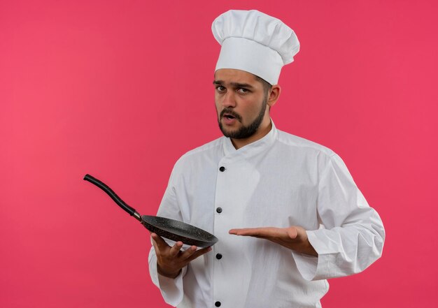 Photo gratuite jeune homme cuisinier impressionné en uniforme de chef tenant et pointant avec la main sur une poêle à frire isolée sur un mur rose