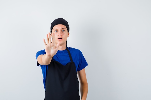 Jeune homme cuisinier montrant un geste d'arrêt en t-shirt, tablier et semblant sérieux, vue de face.