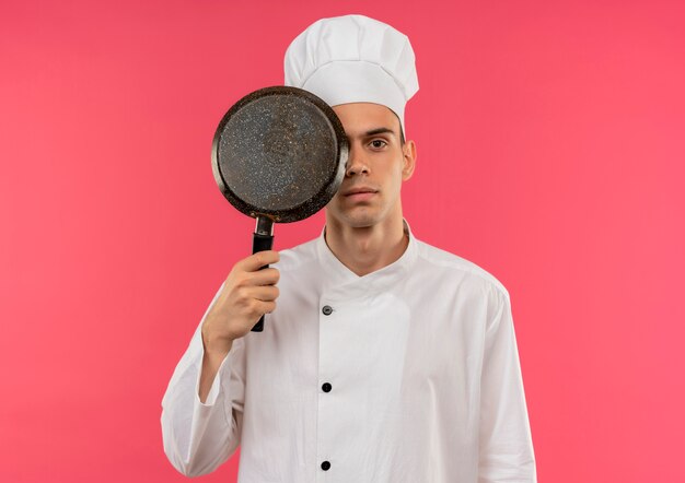 Jeune homme cuisinier portant l'uniforme de chef couvert oeil avec poêle à frire sur mur rose isolé