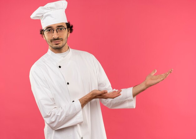 Jeune homme cuisinier portant l'uniforme de chef et des lunettes points avec les mains à l'autre