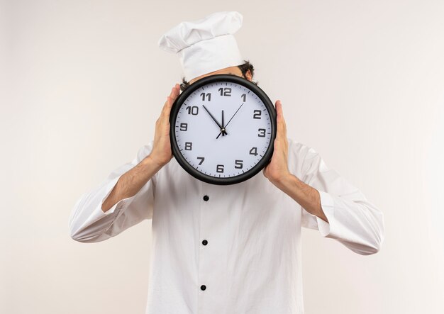 Jeune homme cuisinier portant l'uniforme de chef et lunettes visage couvert avec horloge murale isolé sur mur blanc